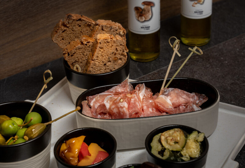 Assorted appetizers with olives, cured meats, bread, and vegetables, accompanied by bottles of mushroom and parmesan oils on a dark wooden surface.