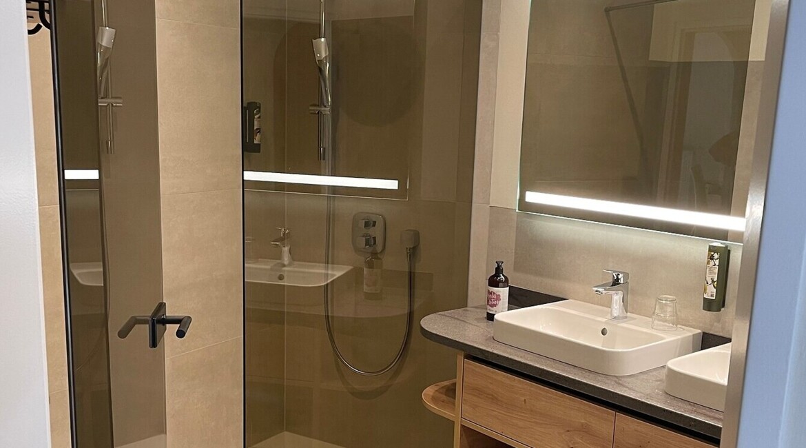 Modern bathroom with a glass shower, wooden vanity, illuminated mirror, and a stool.