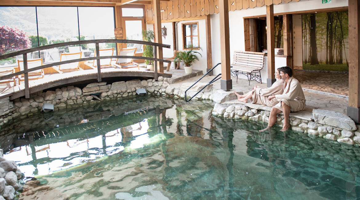 Zwei Personen entspannen am Rand eines Innenpools mit Steinverkleidung in einem gemütlichen Spa-Bereich.