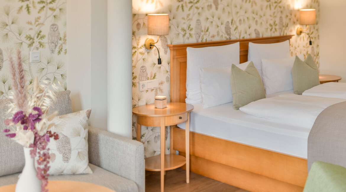 Bright, cozy hotel room with floral wallpaper, wooden furniture, and a vase of dried flowers, offering a comfortable and inviting ambiance.