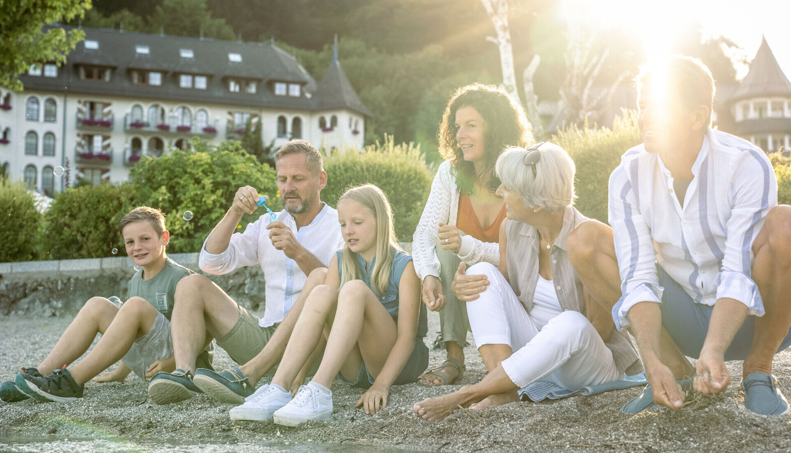 A family enjoying a sunny day by the water, with a scenic hotel and lush greenery in the background.