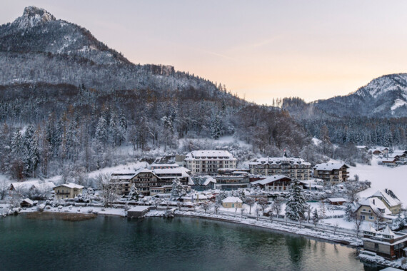 Snow-covered village by a lake with forested mountains in the background at sunrise.