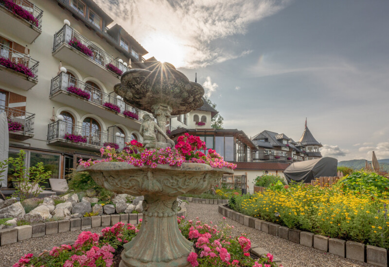 Elegant hotel with balconies adorned with vibrant flowers, a decorative fountain, and a well-maintained garden under a clear sky.