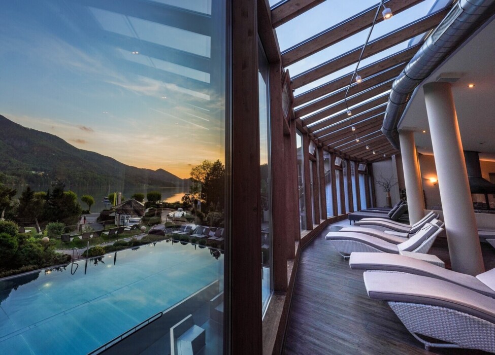 Glass-walled lounge with sun chairs overlooking a scenic lake and mountains at sunset.