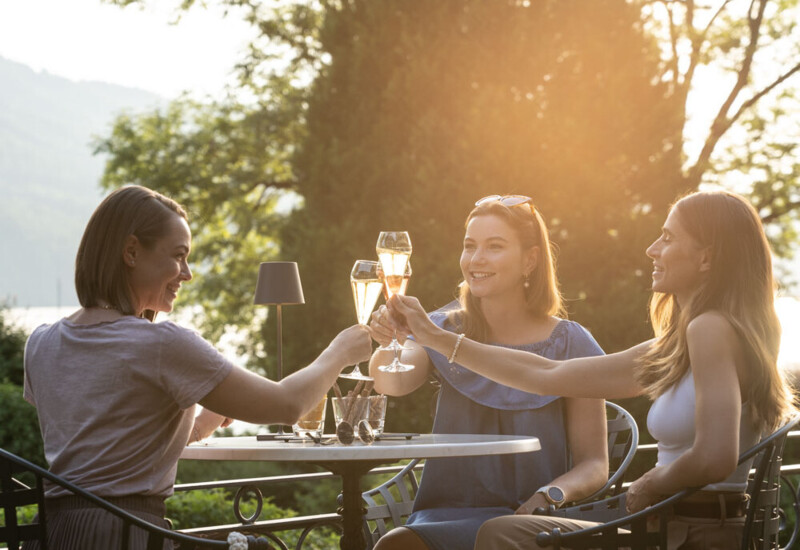 Drei Frauen stoßen auf einer Terrasse bei Sonnenuntergang mit Getränken an.