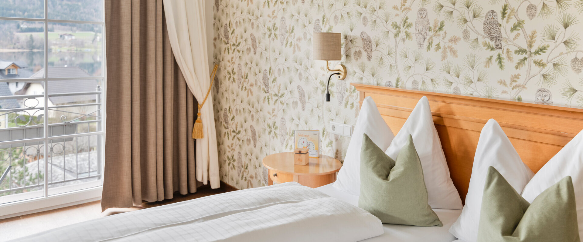 A cozy hotel room with floral and owl wallpaper, a double bed with green pillows, and a scenic view of the lake and mountains through a large window.