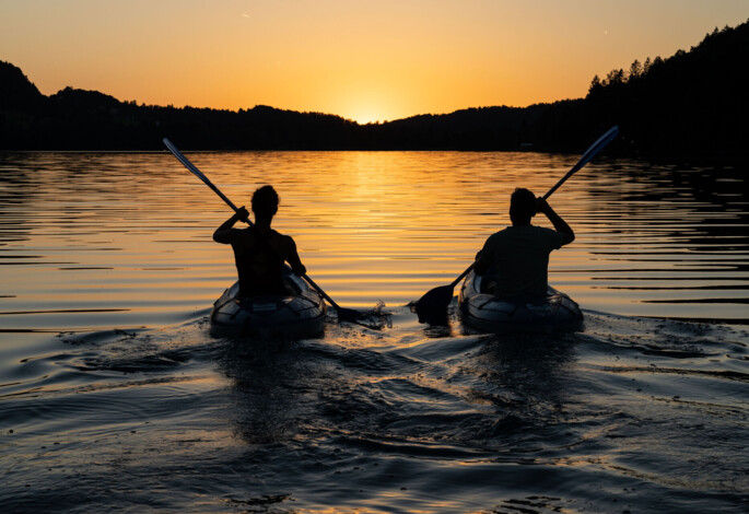 Zwei Personen paddeln bei Sonnenuntergang auf einem See in Kanus.