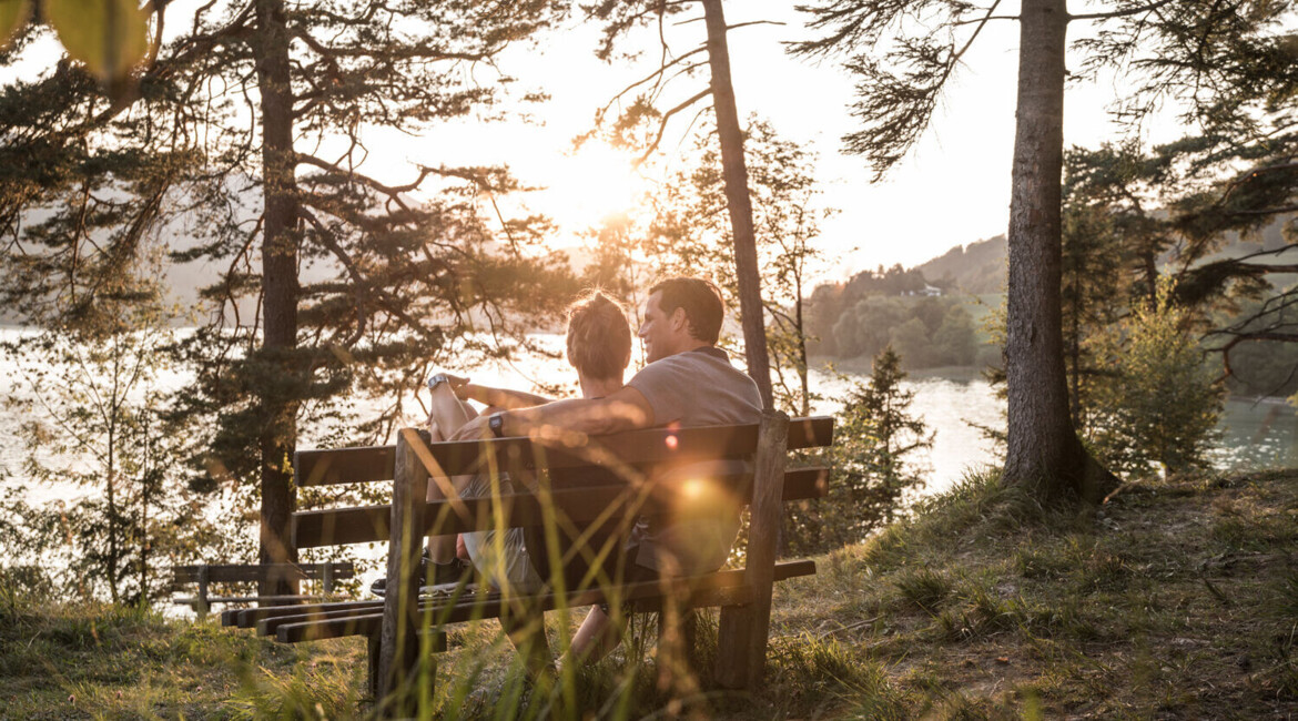 A couple sits on a bench enjoying a serene lake view surrounded by trees during sunset.