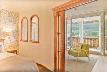 Elegant hotel room with a cozy armchair and balcony offering a scenic view of lush, green hills.