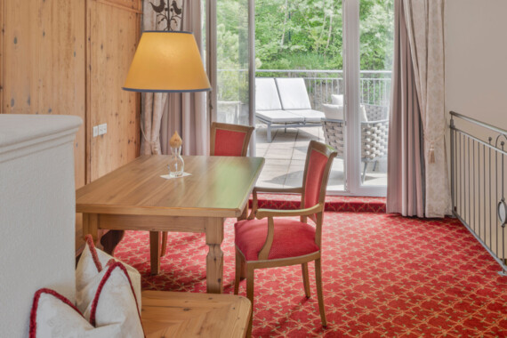 Cozy room with wooden table and red chairs, overlooking a lush balcony with sun loungers through glass doors.