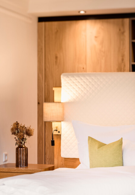 Elegant hotel room with white bedding, a green pillow, and a bedside table with dried flowers in a vase, creating a cozy atmosphere.