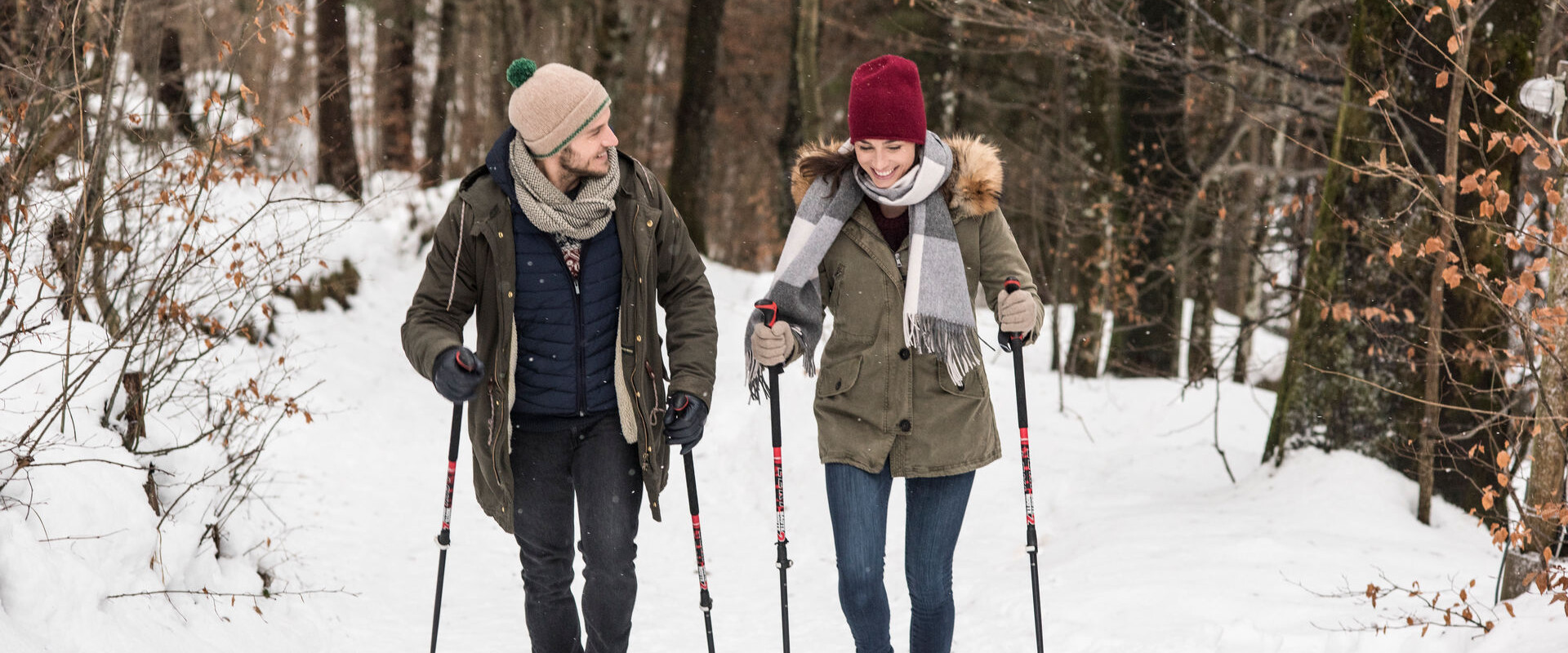 Zwei Personen genießen einen Winterspaziergang mit Stöcken im verschneiten Wald, warm gekleidet mit Mützen und Schals.