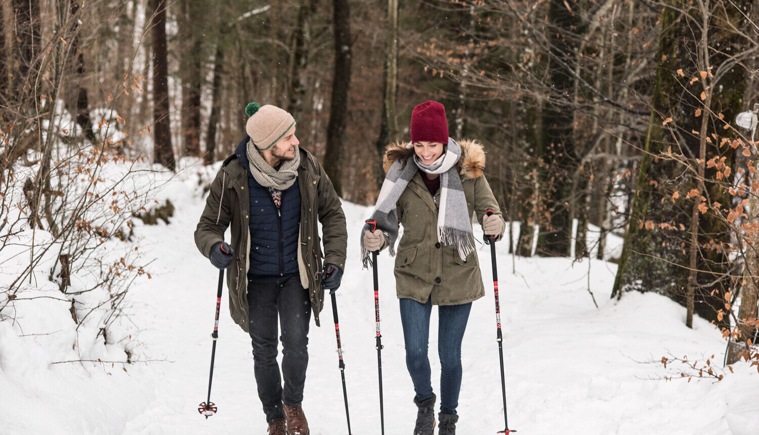A couple enjoys a winter hike through a snowy forest, using trekking poles for support.