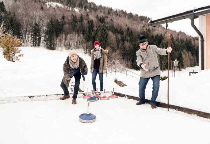 Eine Gruppe von Menschen spielt Eisstockschießen im Schnee vor einer bewaldeten Berglandschaft.