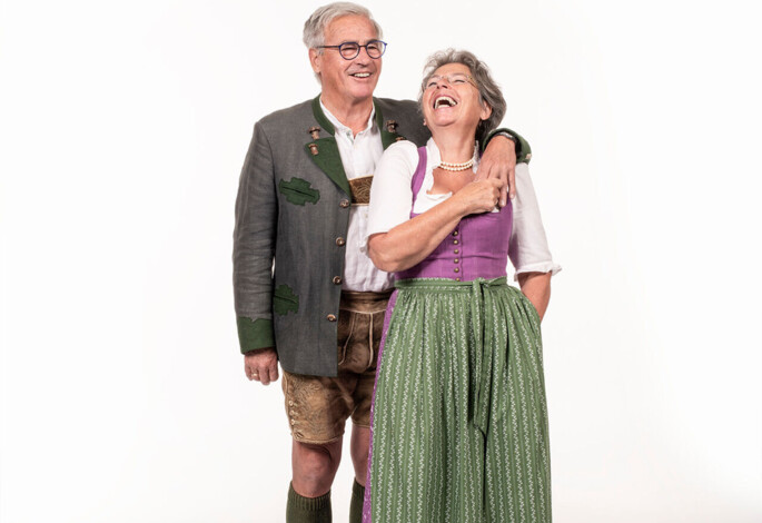 Ein älteres Paar in traditioneller Tracht lacht zusammen vor einem weißen Hintergrund.