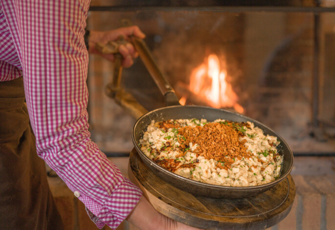 A person in a pink checkered shirt holds a pan of traditional Kasnock'n topped with crispy onions, in front of a warm fireplace.