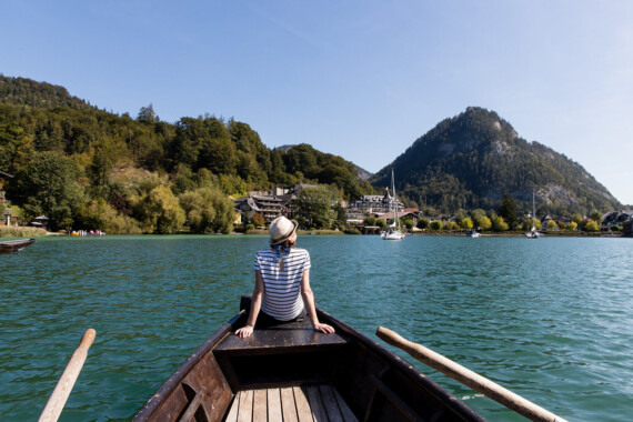 A person in a boat on Fuschlsee lake, facing a mountain and surrounded by trees and buildings, near Waldhof Fuschlsee Resort.