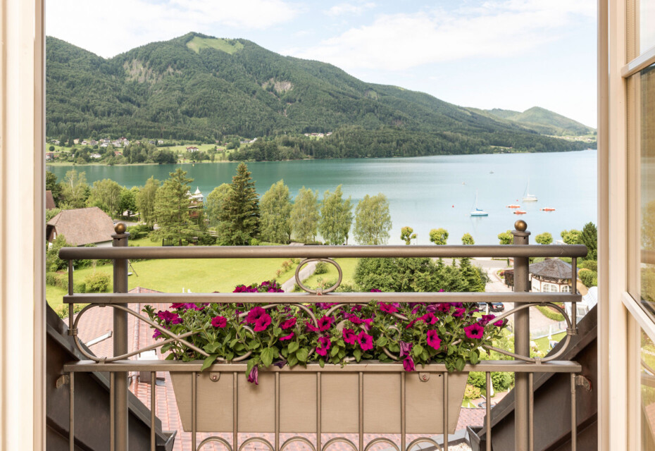 Blick von einem Balkon mit Blumen auf den malerischen Fuschlsee und die umliegenden grünen Berge.