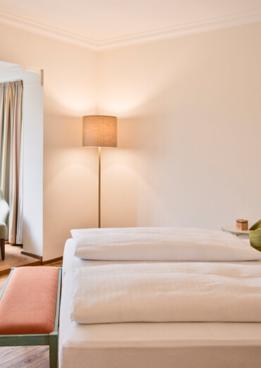 Bright hotel room with a cozy double bed, green pillows, seating area, and large windows overlooking trees.