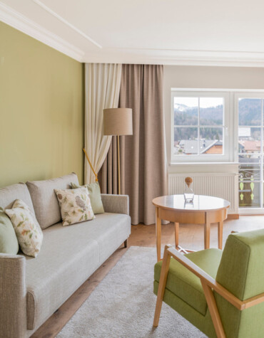 Bright living room with green accents, cozy sofa, armchair, and wooden table by a window offering scenic views.