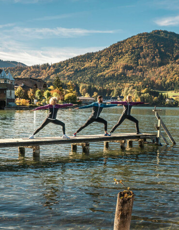 Drei Personen machen Yoga auf einem Steg am Fuschlsee, umgeben von herbstlichen Bergen und klarem Wasser.