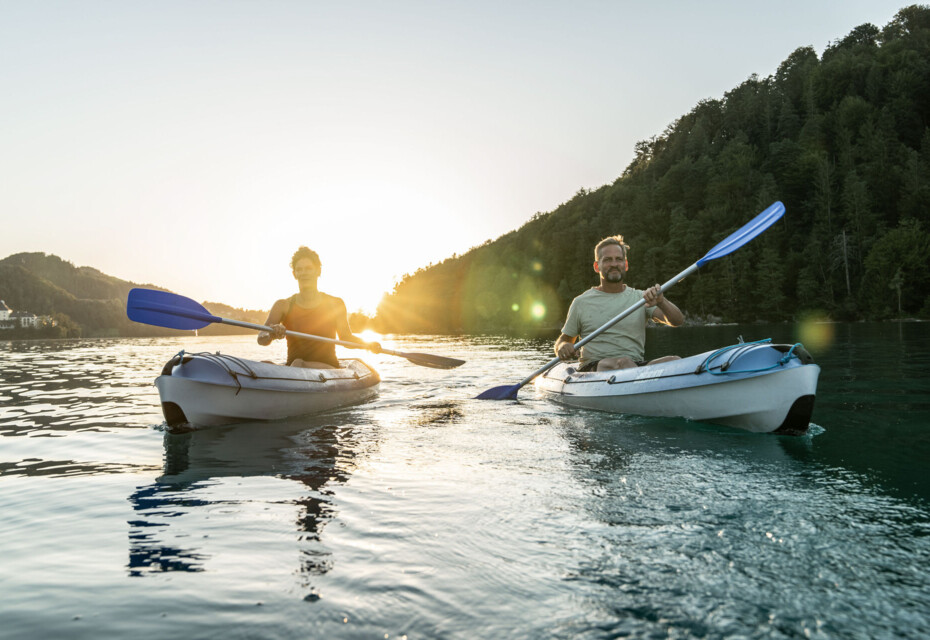 Two people kayaking on a serene lake at sunset, surrounded by lush greenery.