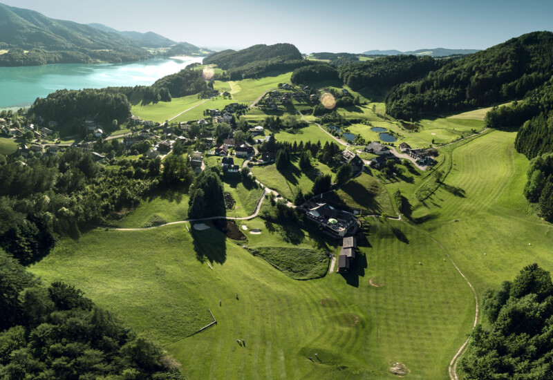Aerial view of a scenic, lush green landscape with a village and lake surrounded by forested hills.