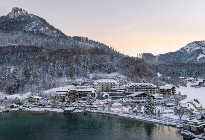 Winterlandschaft mit verschneiten Gebäuden am Fuschlsee, umgeben von bewaldeten Bergen bei Sonnenuntergang.