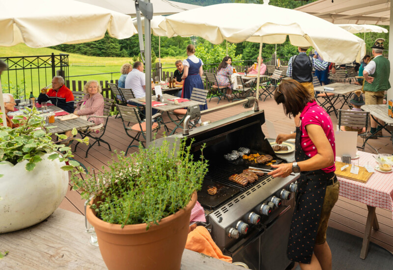 Grillköchin bereitet Fleisch für Gäste auf einer sonnigen Terrasse mit Blick auf die Natur zu. Menschen genießen Essen und Gesellschaft an Tischen.