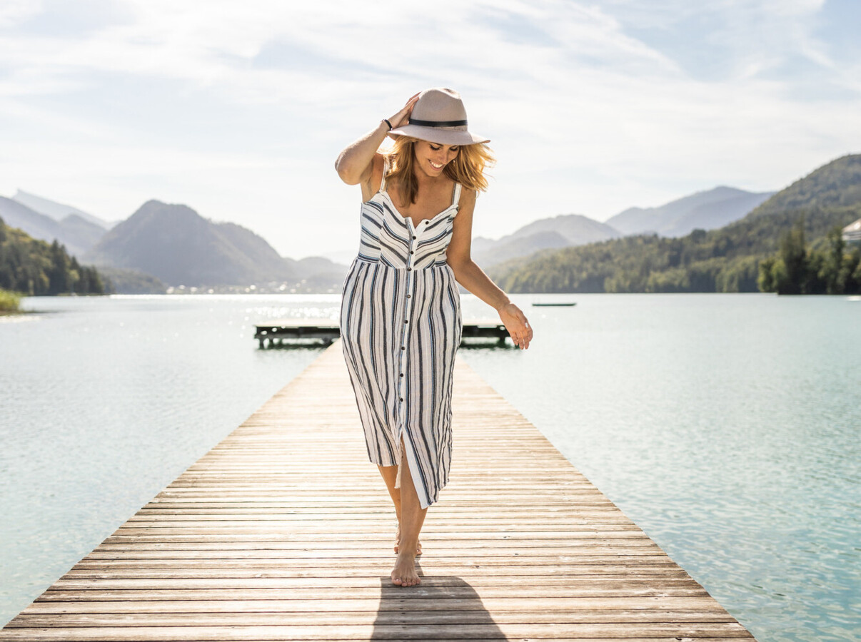A woman in a striped dress and hat walks on a wooden pier over a scenic lake, surrounded by lush mountains under a clear sky.