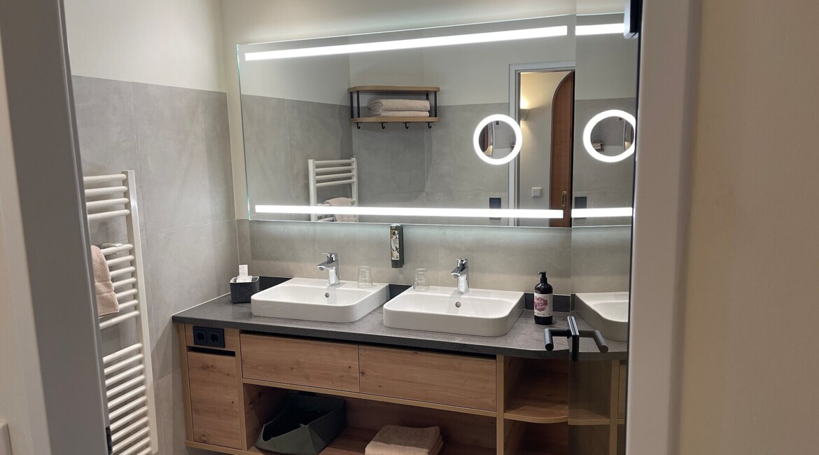 Modernes Badezimmer mit Doppelwaschbecken, großen beleuchteten Spiegeln und Holzunterschrank im Hotel Waldhof Fuschlsee.