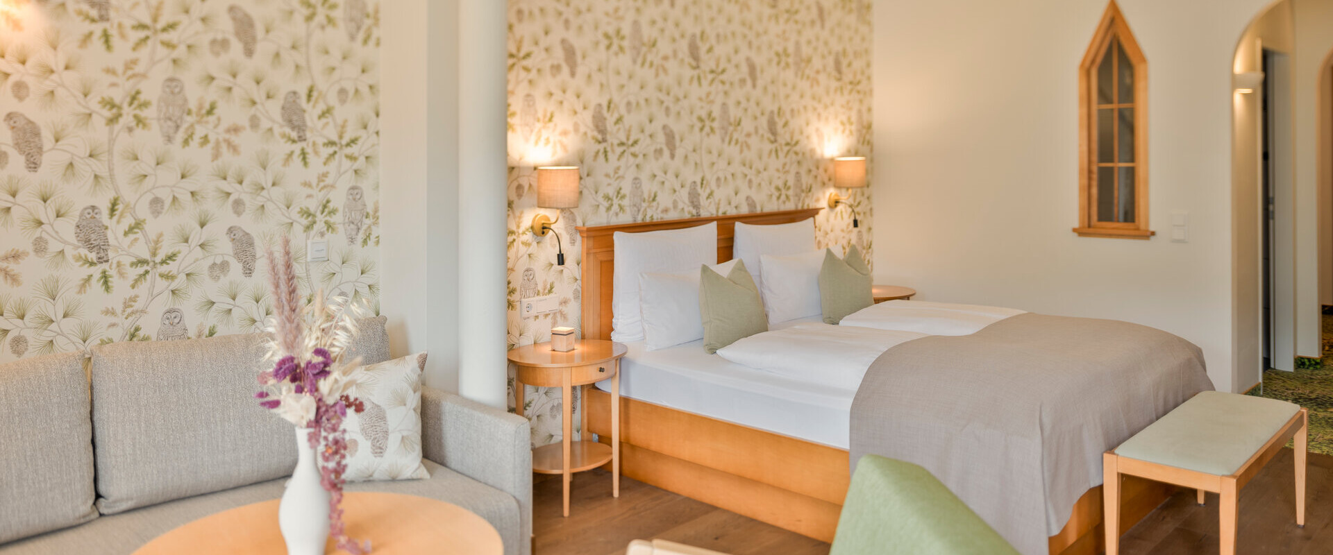 Bright, cozy hotel room with floral wallpaper, a double bed, sofa, and armchair. Soft lighting creates a welcoming atmosphere.