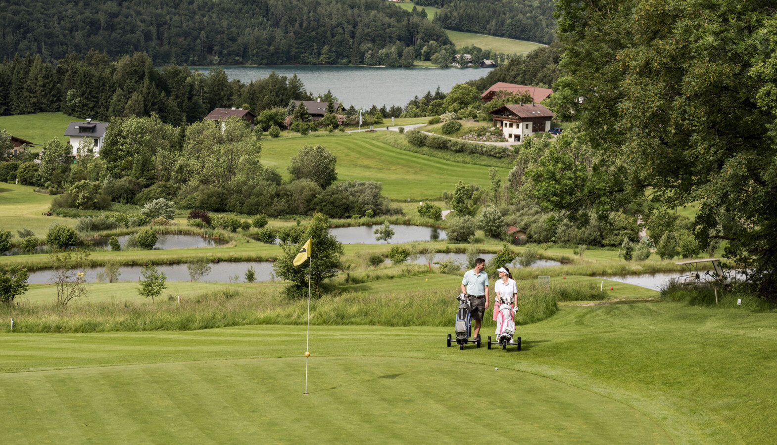 Two people walking on a golf course near Waldhof Fuschlsee Resort, with a flag in the foreground and a lake in the background.
