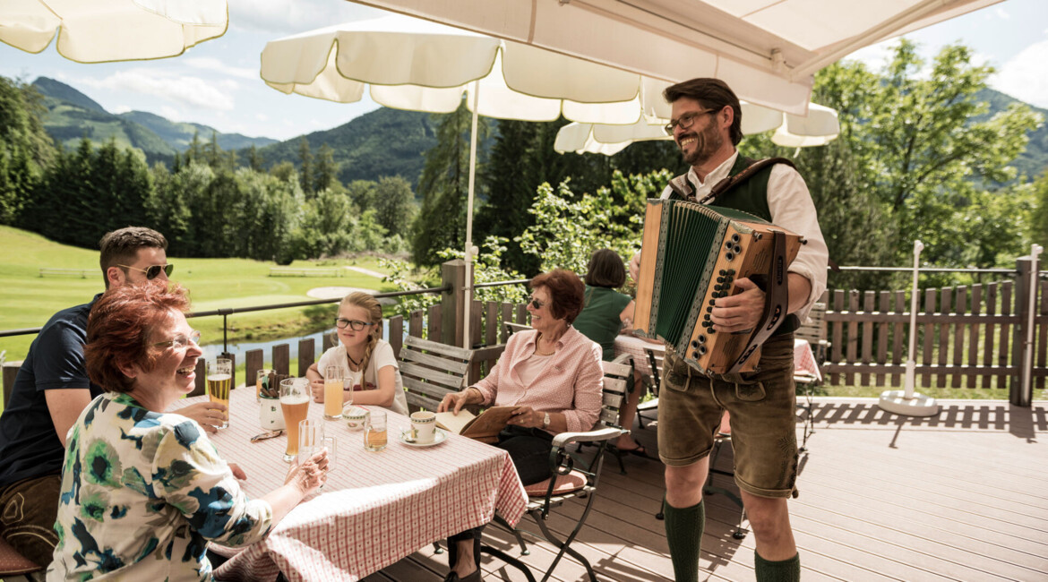 Eine Familie genießt Getränke auf einer sonnigen Terrasse, während ein Musiker mit Akkordeon spielt, umgeben von idyllischer Berglandschaft.