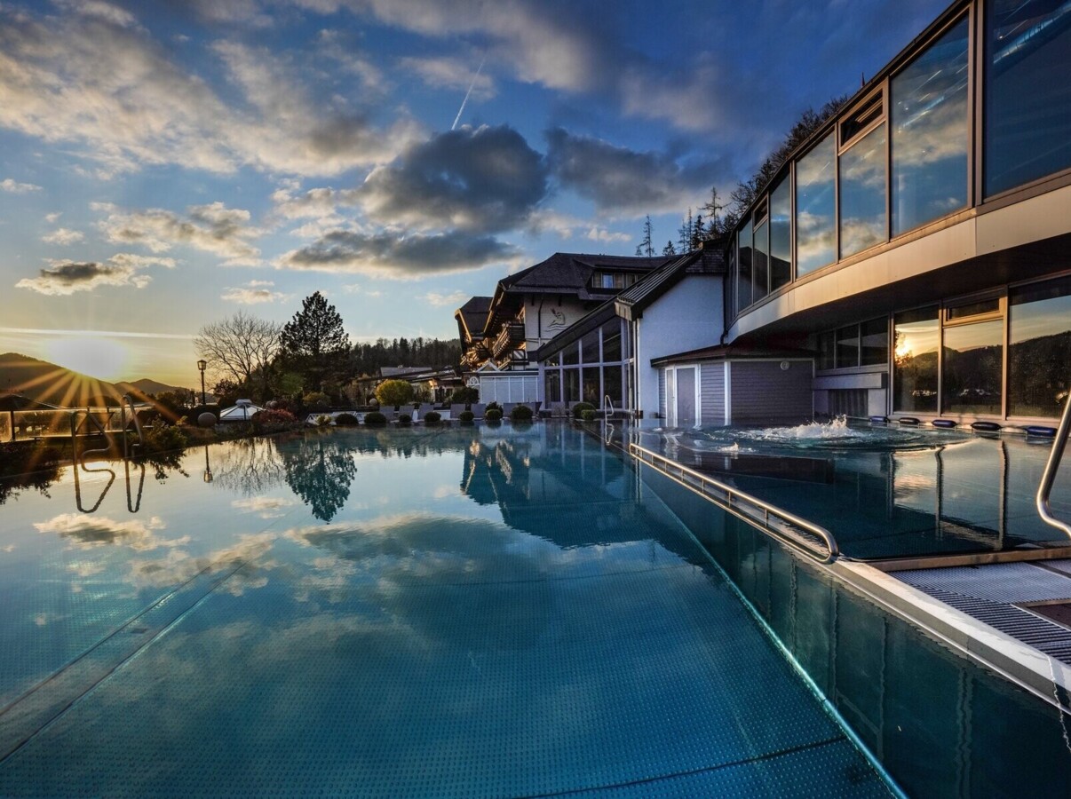 Outdoor pool reflecting the sunset and clouds, with a modern building and natural landscape in the background.