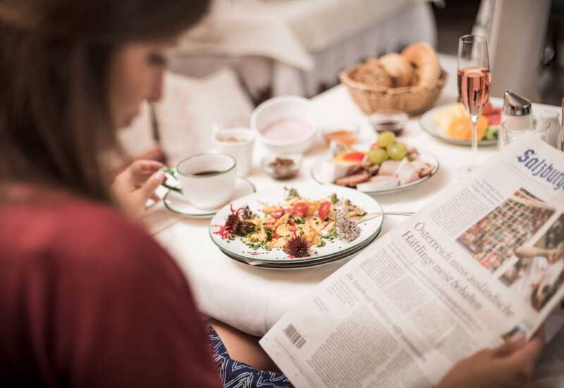 A person enjoys breakfast with a newspaper, featuring a plate of scrambled eggs and a variety of foods on a table.