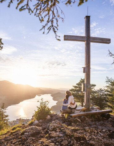 A couple sits by a wooden cross on a mountain with a view of a lake and sunset.