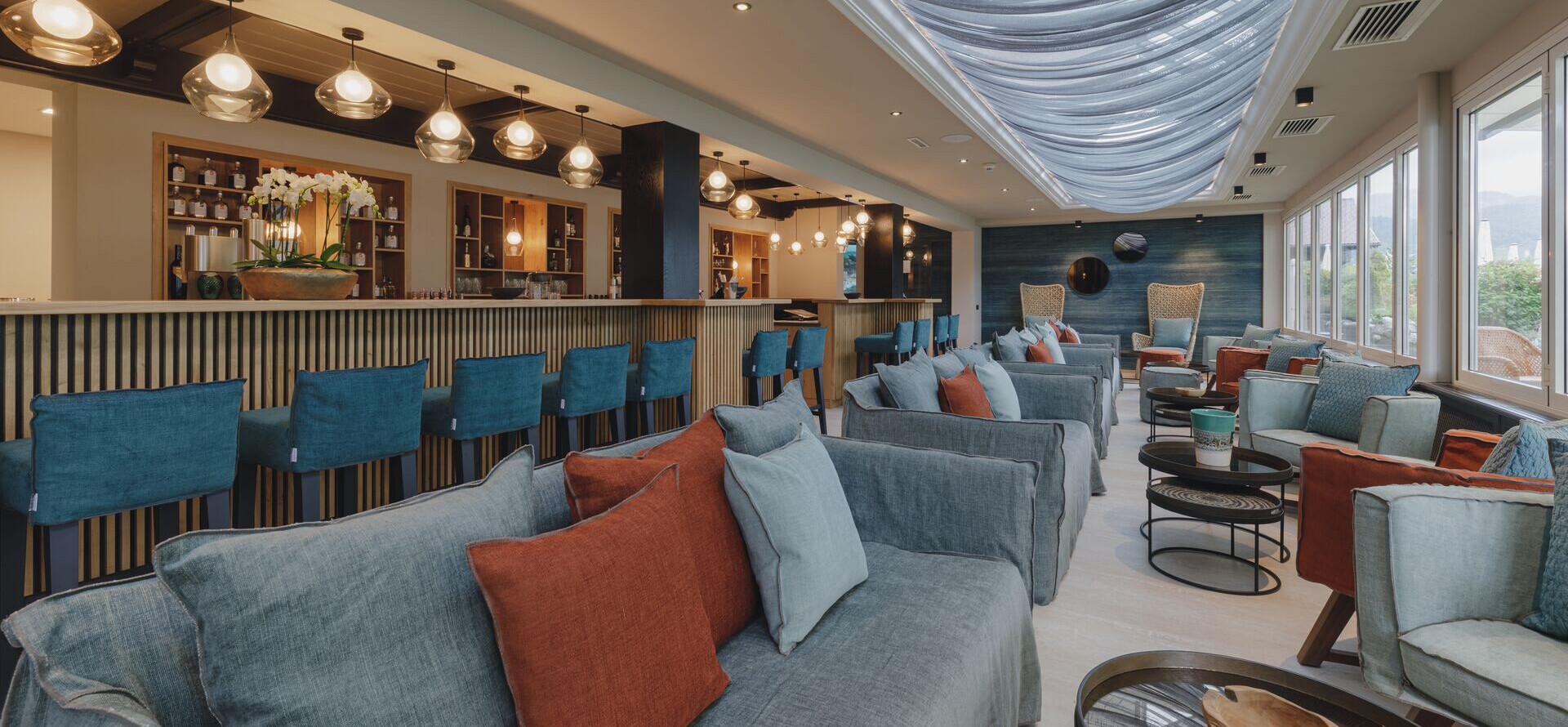Modern lounge area with cozy seating, colorful cushions, and a stylish bar under a draped ceiling, illuminated by elegant pendant lights.