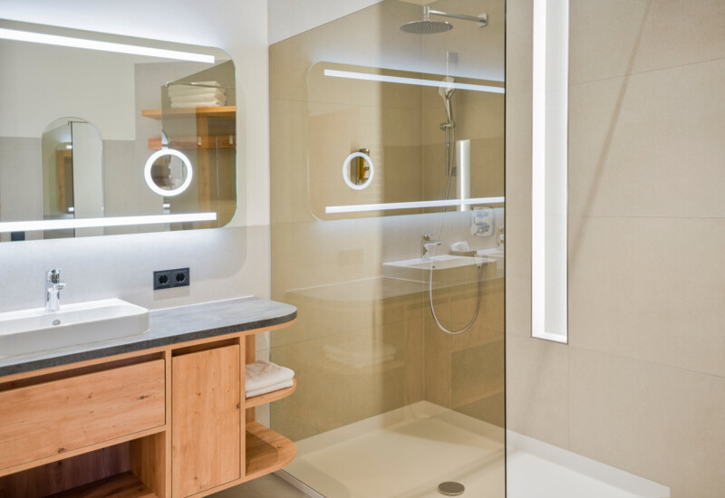 Modernes Badezimmer mit großer Dusche und Spiegeln. Holzmöbel und beleuchtete Spiegel sorgen für ein elegantes Ambiente.