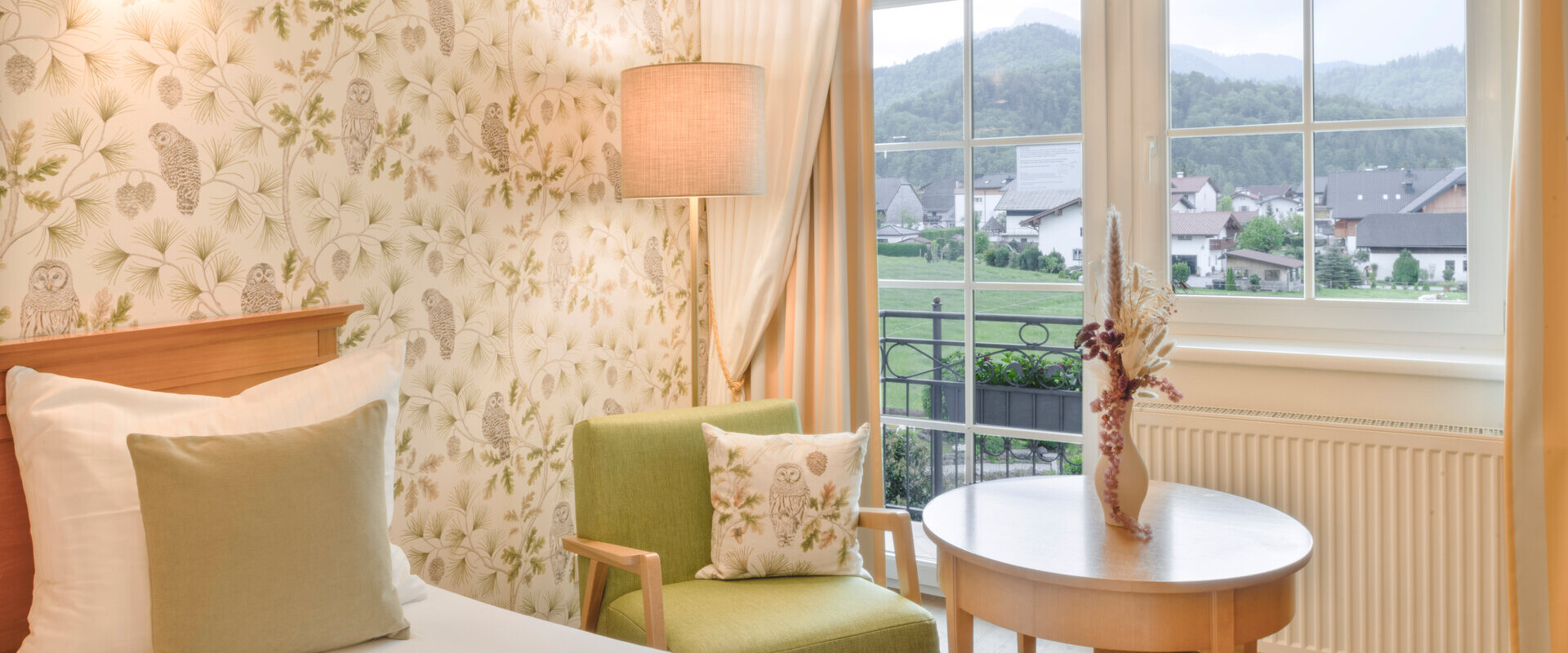 Hotel room with floral wallpaper, a green chair, and a view of the countryside at Waldhof Fuschlsee Resort.