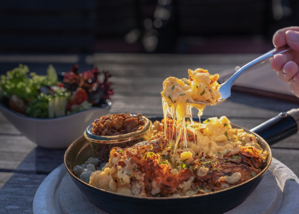 Käsespätzle mit geschmolzenem Käse im Pfännchen, serviert mit einem frischen Salat im Hintergrund auf einem Tisch.