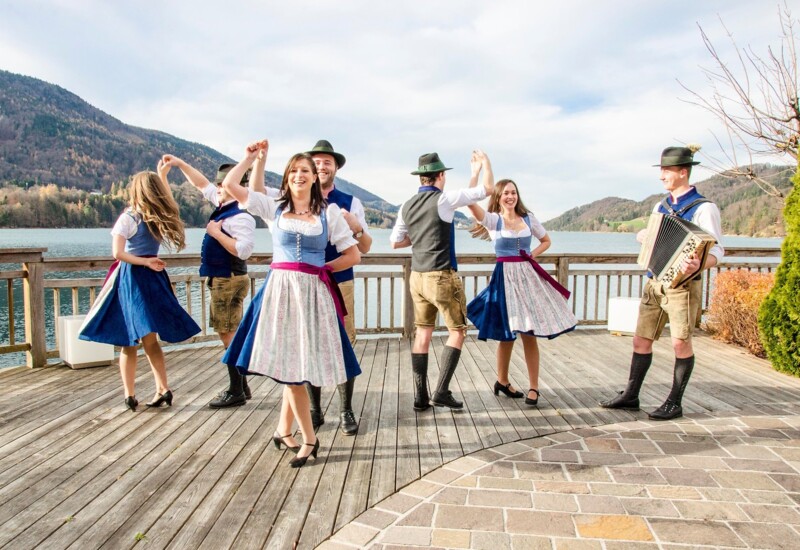 Tänzer in traditioneller Tracht tanzen auf einem Holzboden vor einer Berg- und Seelandschaft.