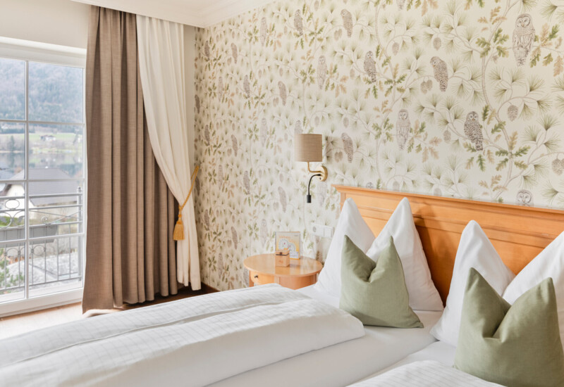 A cozy hotel room with floral and owl wallpaper, a double bed with green pillows, and a scenic view of the lake and mountains through a large window.