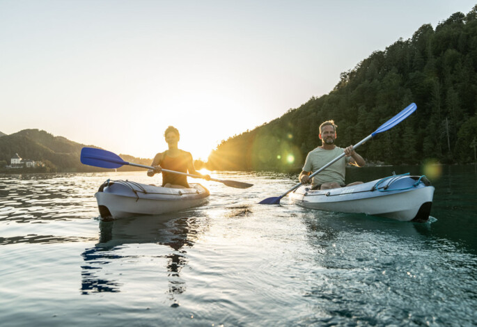Two people kayaking on a calm lake at sunset, surrounded by lush, scenic forested hills.