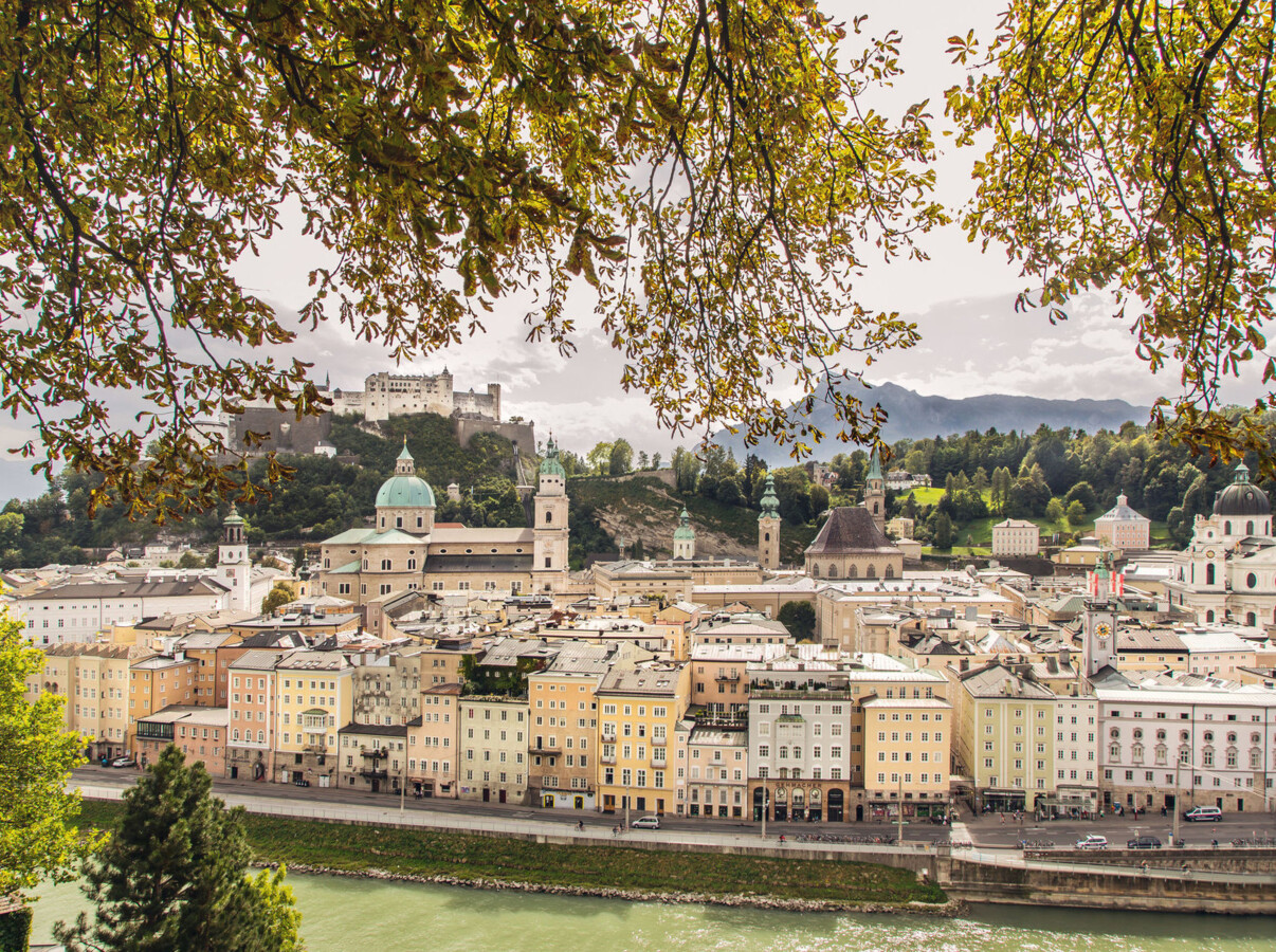 View of Salzburg cityscape with fortress and cathedral, framed by lush tree branches, overlooking the scenic river.