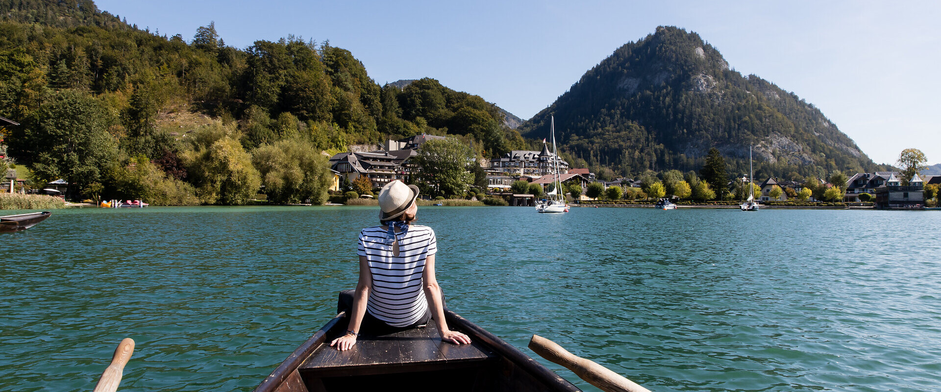 A person in a boat on Fuschlsee lake, facing a mountain and surrounded by trees and buildings, near Waldhof Fuschlsee Resort.