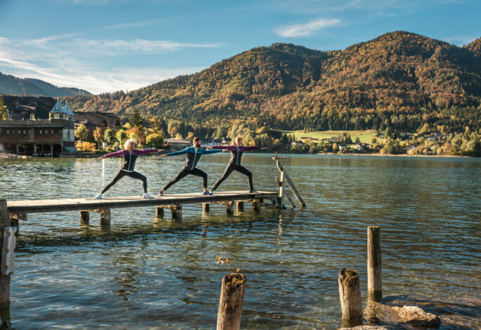 Three people practicing yoga on a wooden dock by lake Fuschl, surrounded by scenic mountains in autumn.