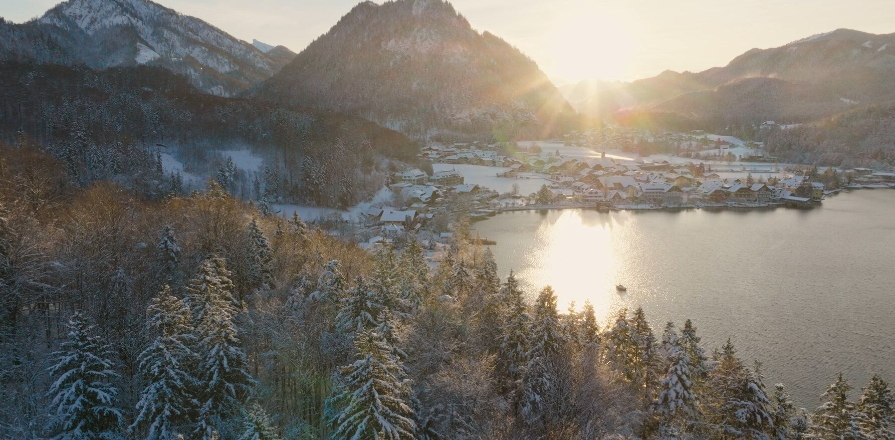 Snow-covered landscape with sunlit mountains and a shimmering lake, surrounded by frosted trees and a quaint village in the distance.