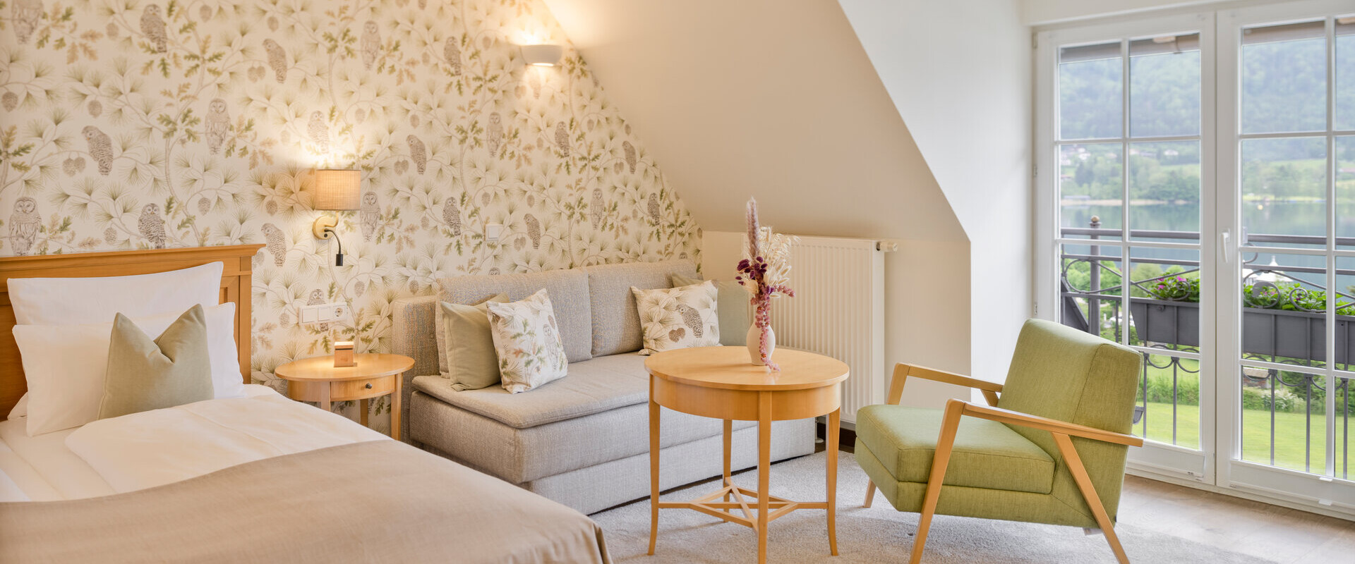 Elegant hotel room with floral wallpaper, cozy bed, sofa, and armchair, overlooking a scenic landscape through large windows.