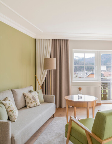 Bright hotel room with green accents, cozy sofa, armchair, and wooden table by a window offering scenic views.